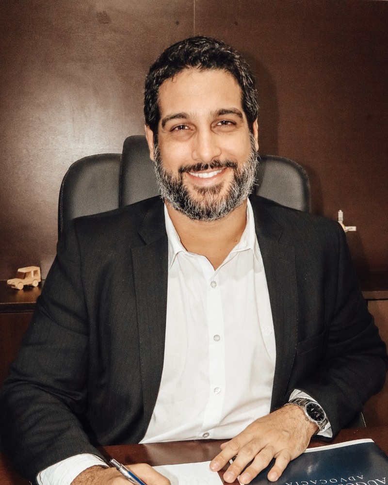 O advogado Augusto Neto, de terno escuro e camisa branca, sentado à mesa do escritório, olhando para a câmera
