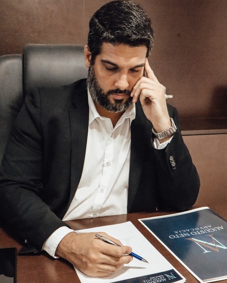 O advogado Augusto Neto à mesa do escritório, de terno escuro, escrevendo em um documento